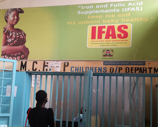 Woman looks up at sign in Kenyan hospital that reads "Iron and Folic Acid Supplements (IFAS) keep me and my unborn baby healthy"