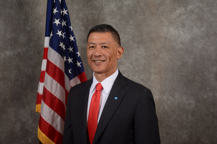 Asian man in his mid-fifties sits in front of an American flag. He is wearing a black suit with a red tie.