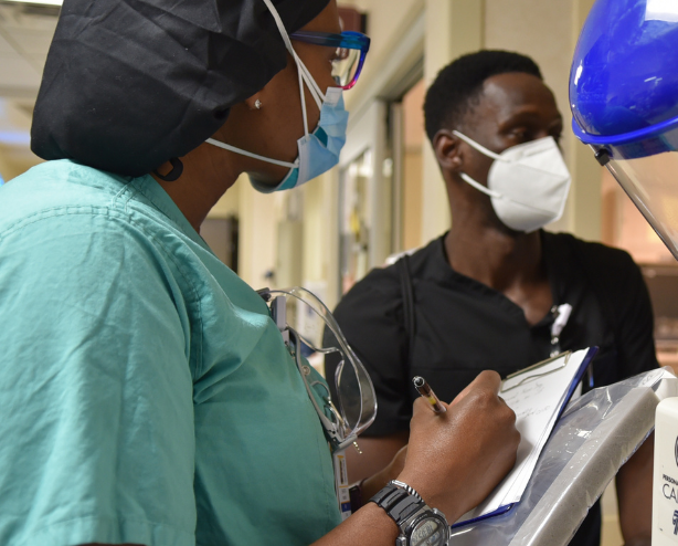 Black woman in medical scrubs and mask holds a clip board and speaks with a Black man