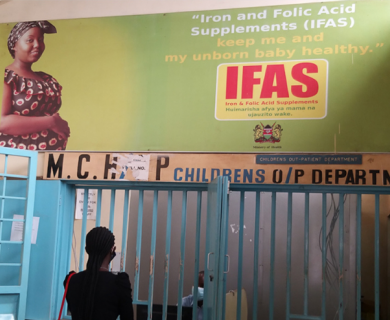 Woman looks up at sign in Kenyan hospital that reads "Iron and Folic Acid Supplements (IFAS) keep me and my unborn baby healthy"