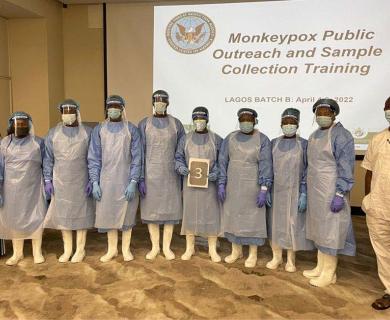 A group of students in personal protective equipment stand in front of a training presentation