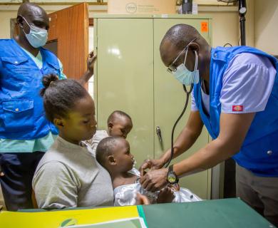 Two masked health workers examine a young girl sitting on her mother's lap.