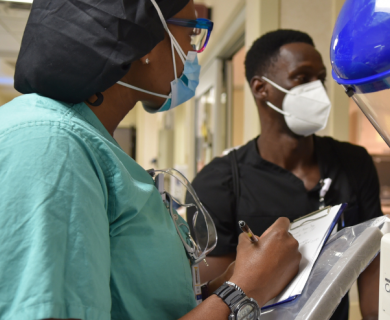 Black woman in medical scrubs and mask holds a clip board and speaks with a Black man
