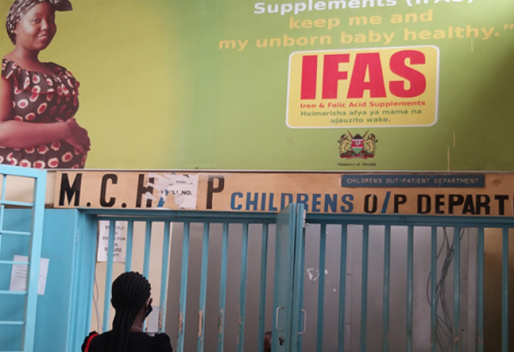 Woman looks up at sign in Kenyan hospital that reads "Iron and Folic Acid Supplements (IFAS) keep me and my unborn baby healthy"