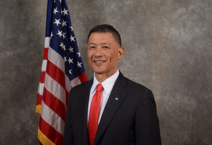 Asian man in his mid-fifties sits in front of an American flag. He is wearing a black suit with a red tie.