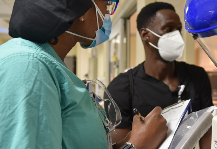 Black woman in medical scrubs and mask holds a clip board and speaks with a Black man