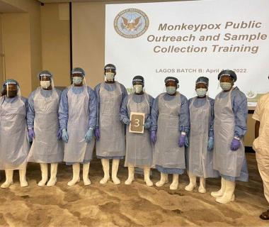 A group of students in personal protective equipment stand in front of a training presentation