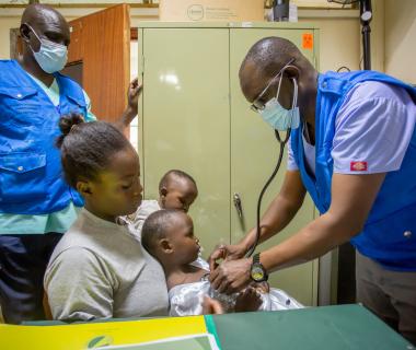 Two masked health workers examine a young girl sitting on her mother's lap.