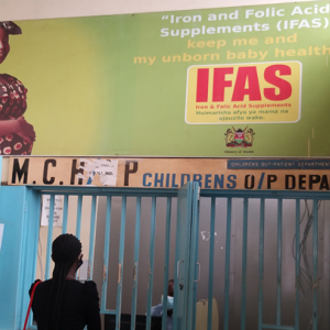 Woman looks up at sign in Kenyan hospital that reads "Iron and Folic Acid Supplements (IFAS) keep me and my unborn baby healthy"